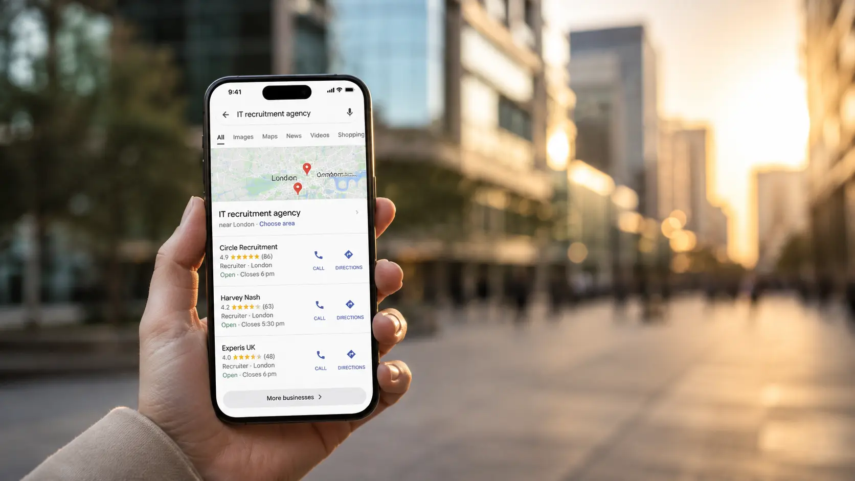 A hand holds a smartphone showing local Google results for “IT recruitment agency” against a blurred urban business district at golden hour.